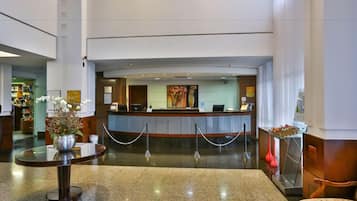 Zona de recepción de hotel con mostrador, área de espera con mesa y sillas, y un jarrón decorativo con flores.