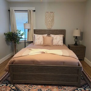 Une chambre à coucher comprenant un lit, une table de chevet, une lampe, une décoration murale en macramé et une plante en pot.