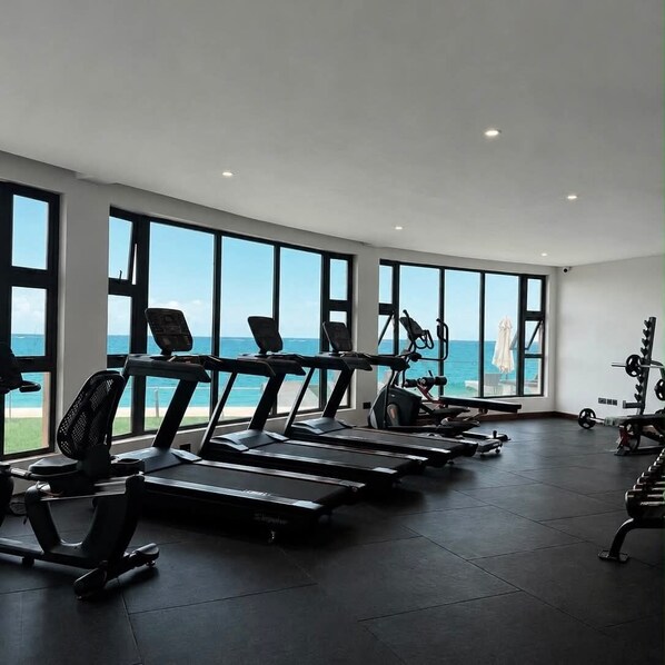Un gimnasio con cintas de correr, bicicletas estáticas y máquinas de pesas, con amplias ventanas y vistas al océano.