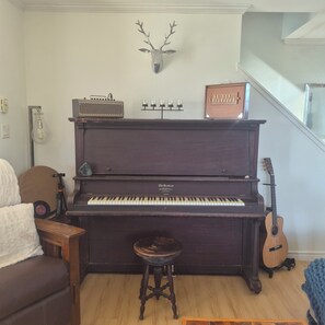 A piano with a stool in front, a wall-mounted deer head, a vintage radio, and a guitar to the right. - Song Sparrow Cottage - Barrel Sauna / Water Views / Short walk to beach (North Rustico)
