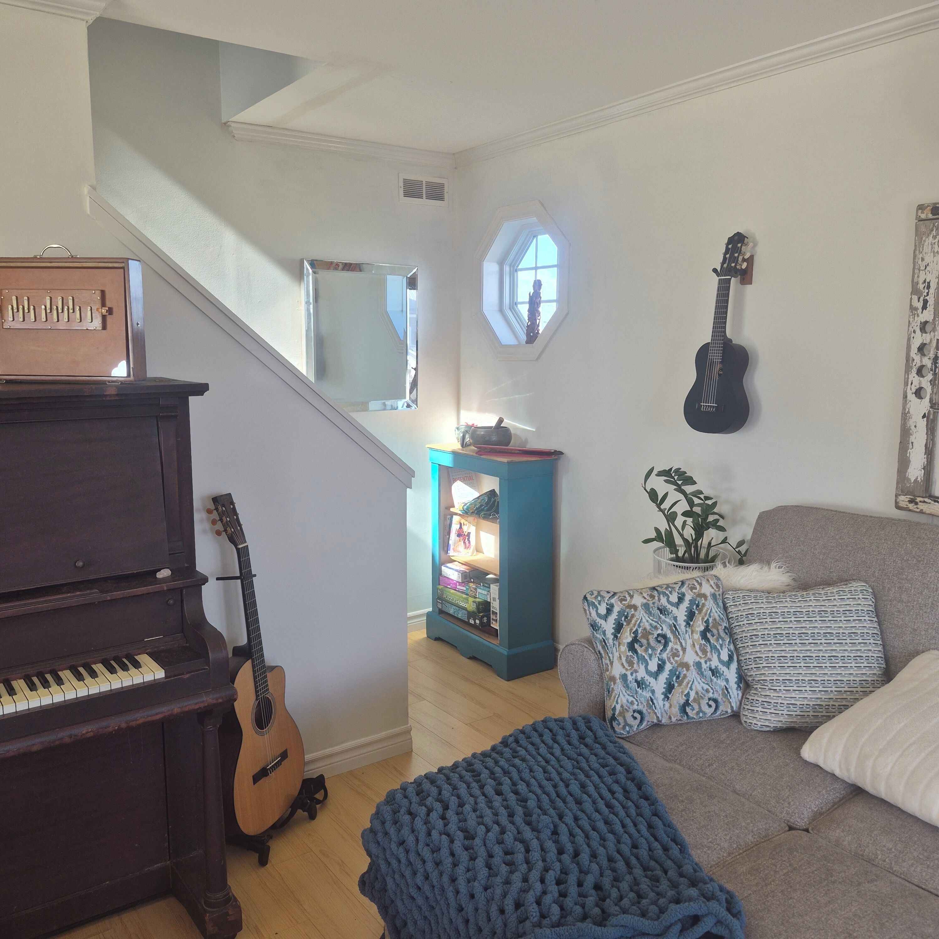 A living room with a piano, a guitar, a blue knitted blanket, and a couch with patterned cushions.