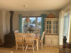 A dining area with a wooden table and chairs, a white cabinet, and a large window with curtains. - Song Sparrow Cottage - Barrel Sauna / Water Views / Short walk to beach (North Rustico)
