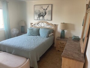 Ein Schlafzimmer mit einem Bett, einem Nachttisch, einer Lampe, einem Spiegel, einem Wandbild von einem Reh und einem Fenster mit Vorhängen.