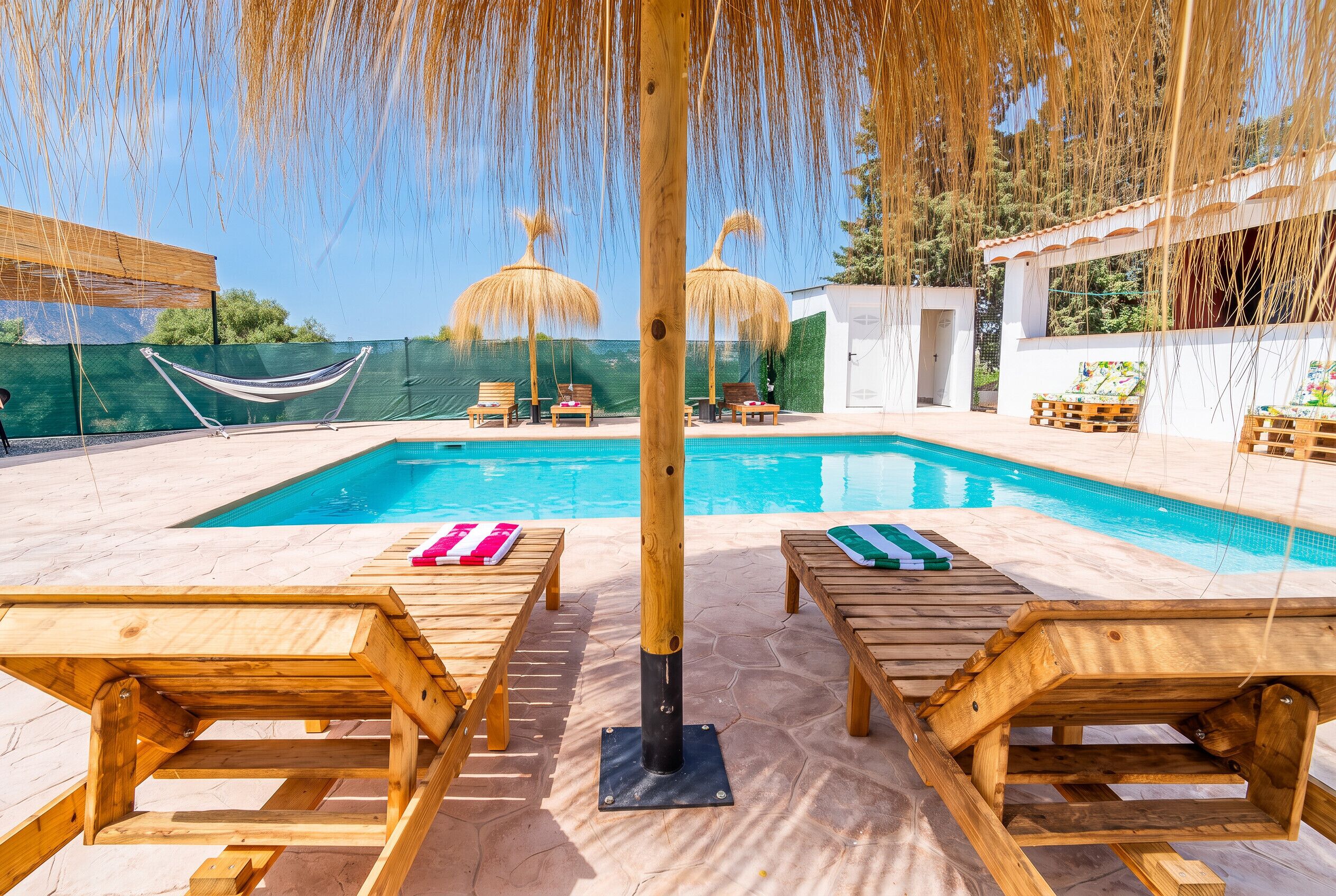 A poolside area with wooden loungers, a hammock, and a small building with a door.