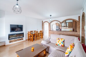Ein Wohnzimmer mit Fernseher, Kamin, Esstisch und einer durch einen Bogen sichtbaren Küche.
