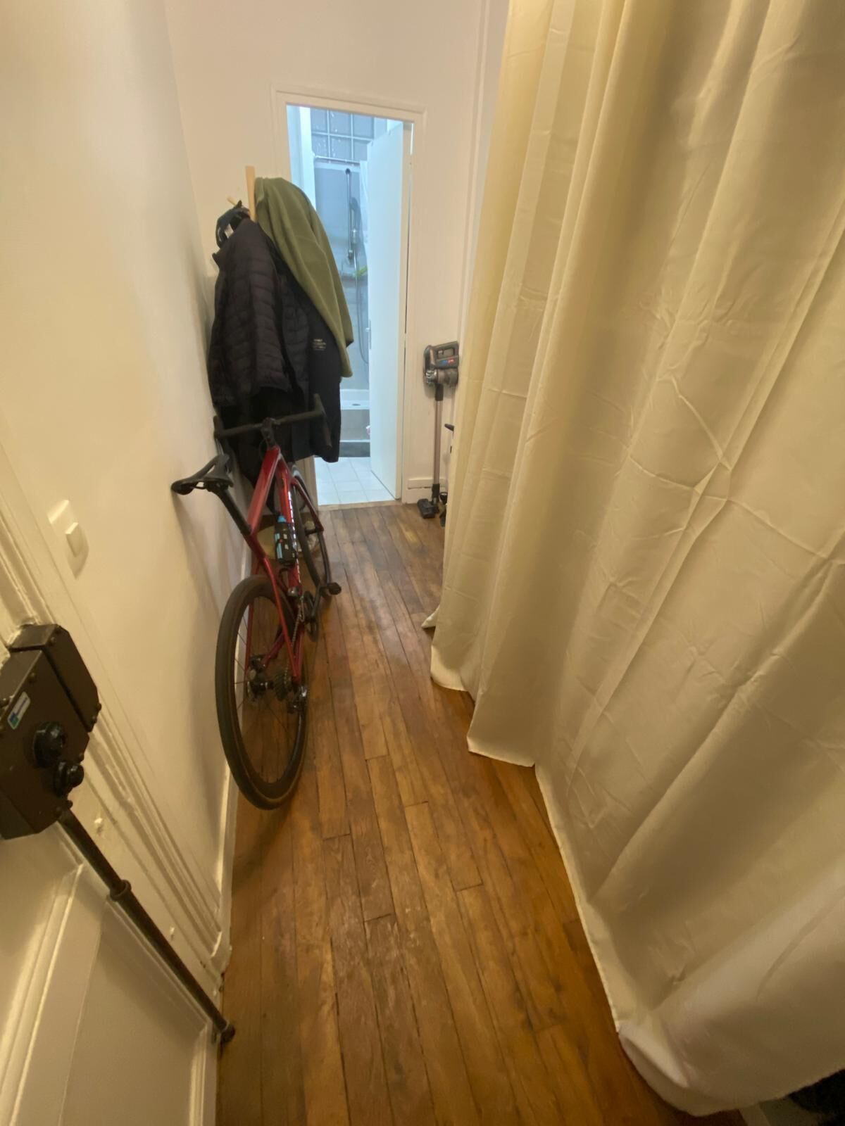 Un pasillo estrecho con una bicicleta apoyada en la pared, una puerta que da al baño y una cortina blanca que cubre parcialmente una ventana.
