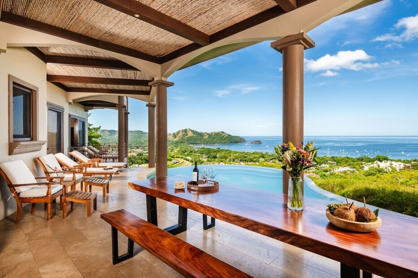 A covered outdoor seating area with a pool, wooden furniture, and a view of the ocean.