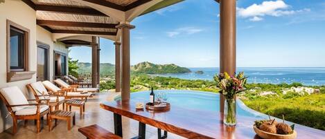 A covered outdoor seating area with a pool, wooden furniture, and a view of the ocean.
