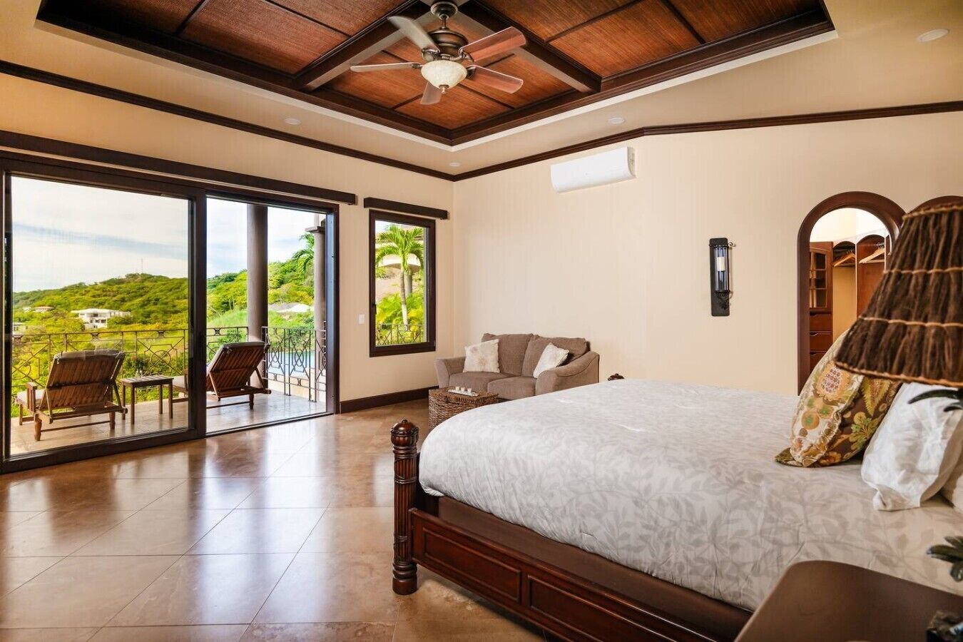 A bedroom with a large bed, a ceiling fan, a view through the glass doors, and a balcony with chairs.