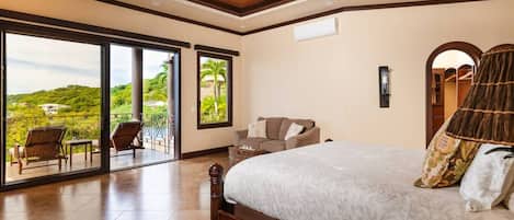 A bedroom with a large bed, a ceiling fan, a view through the glass doors, and a balcony with chairs.