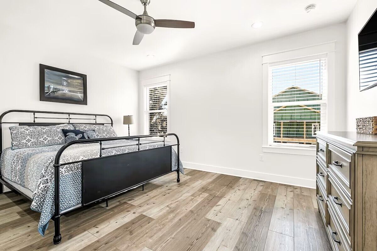 A bedroom with a bed, a ceiling fan, a window with blinds, and a view of a building outside.