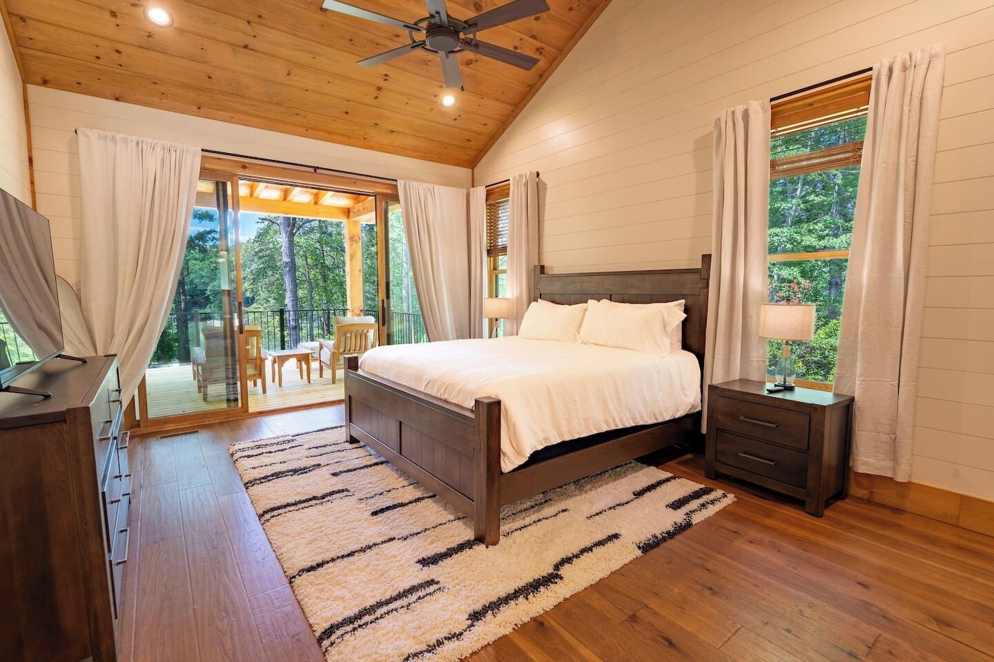 A bedroom with a wooden ceiling, a bed with white bedding, a nightstand, a TV, and a view of the outdoors through a sliding glass door.