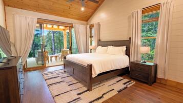A bedroom with a wooden ceiling, a bed with white bedding, a nightstand, a TV, and a view of the outdoors through a sliding glass door.