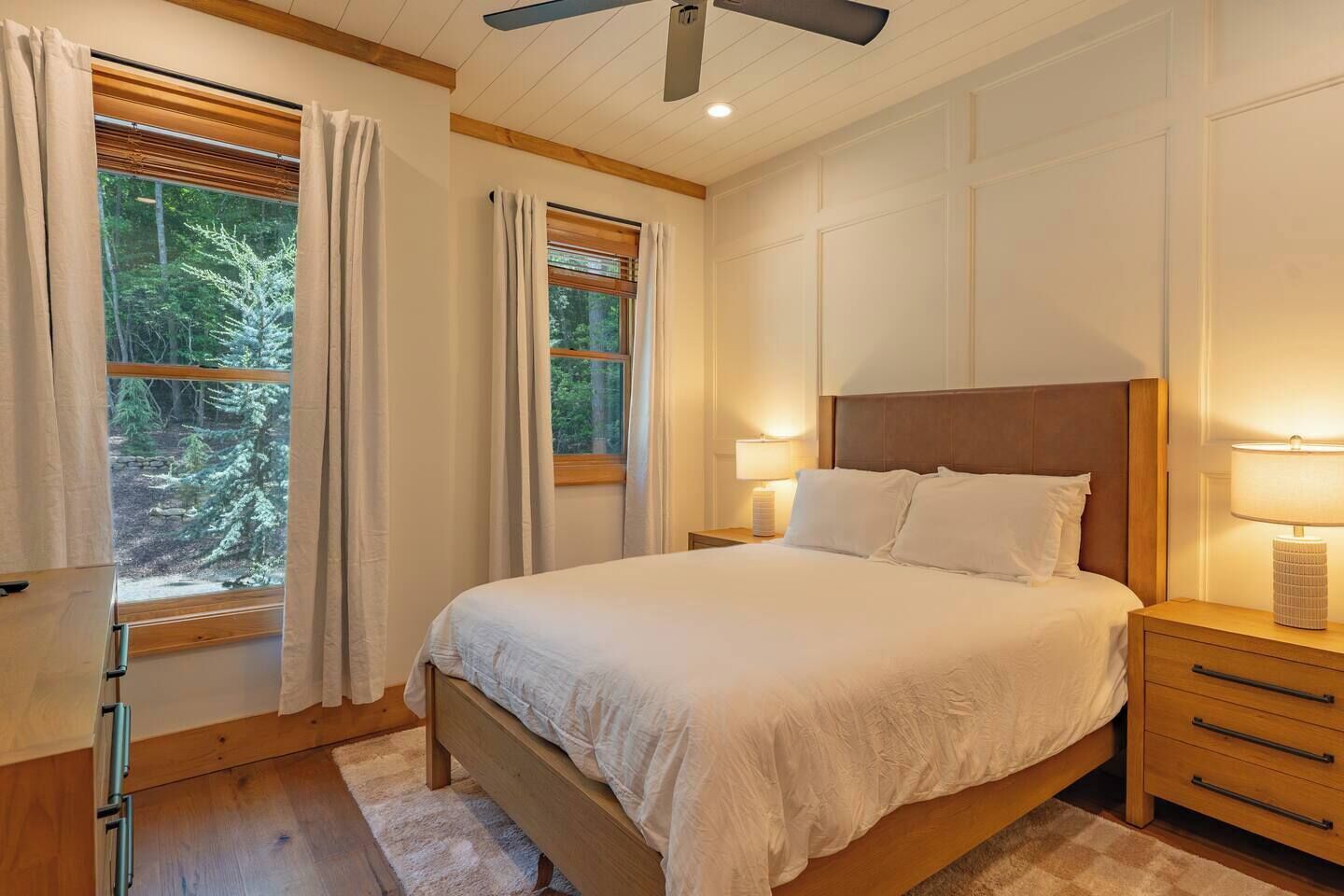 Une chambre à coucher avec un grand lit, deux tables de chevet, un ventilateur de plafond et des fenêtres avec des rideaux.