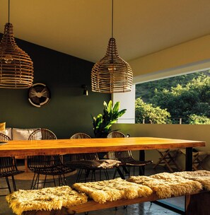 Un espace repas avec une table en bois, des suspensions en fibres tressées et une vue sur la verdure environnante.