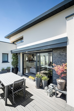 Et moderne hus med en terrasse, spiseområde og et køkken, der kan ses igennem glasdøre.