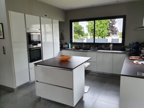 A modern kitchen with white cabinetry, a built-in oven, and a window with a view of trees and buildings. - Holiday Home 'Welcome' with Terraces, Private Garden and Wi-Fi (Le Relecq-Kerhuon)