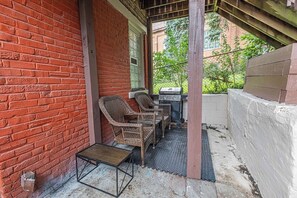 A covered patio area with a brick wall, two wicker chairs, a small table, and a grill. - Mount Washington 4BR Home, Sleeps 10, 2 Kitchens (Pittsburgh)