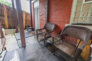 A covered outdoor seating area with wicker chairs and a metal table, adjacent to a red brick wall and a wooden fence. - Mount Washington 4BR Home, Sleeps 10, 2 Kitchens (Pittsburgh)