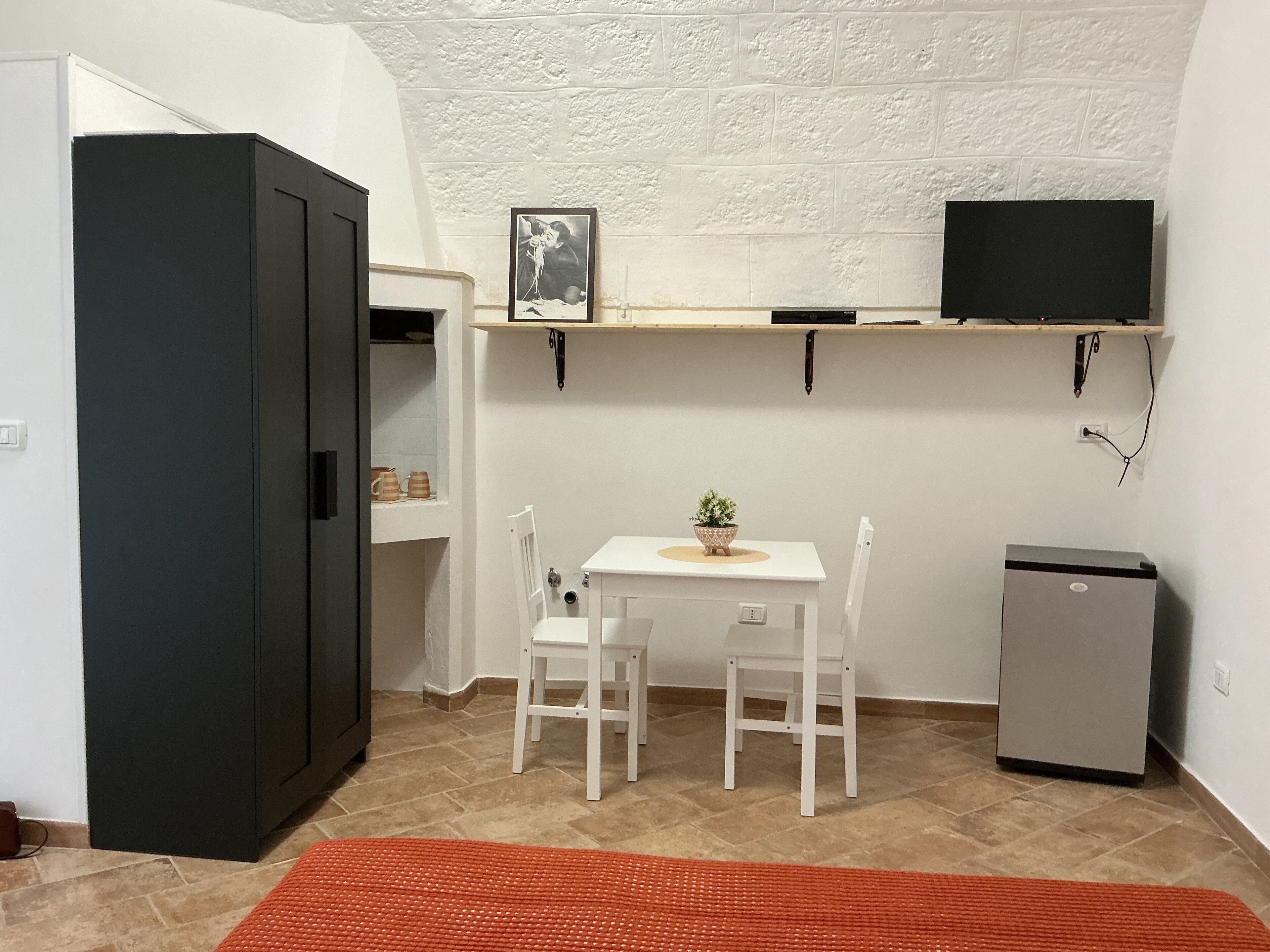 Cocina compacta con refrigerador, una mesita con sillas, una planta en maceta y un televisor montado.