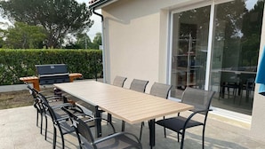 An outdoor patio with a rectangular table and six chairs, a barbecue grill, and a glass door leading to a covered area. - New home in the heart of the Médoc—4 bedrooms, garden, beaches 20 minutes away (Lesparre-Médoc)
