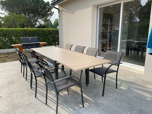 Eine Außenpatio mit einem rechteckigen Tisch und sechs Stühlen, einem Grill und einer Glasdoor, die zu einem überdachten Bereich führt.