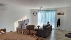 A modern living room with a dining area, a sofa, a television, and a clock on the wall. - New home in the heart of the Médoc—4 bedrooms, garden, beaches 20 minutes away (Lesparre-Médoc)