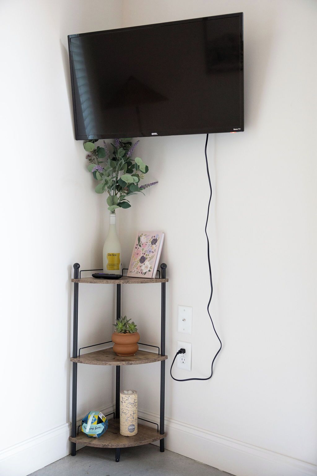 A wall-mounted TV above a metal shelf unit with a potted plant, a picture frame, and a remote control.