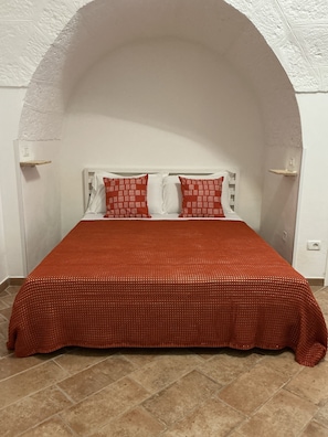 Un letto con una coperta rossa dalla trama particolare e due cuscini con motivo rosso e bianco, posto di fronte a una parete bianca con una nicchia ad arco.