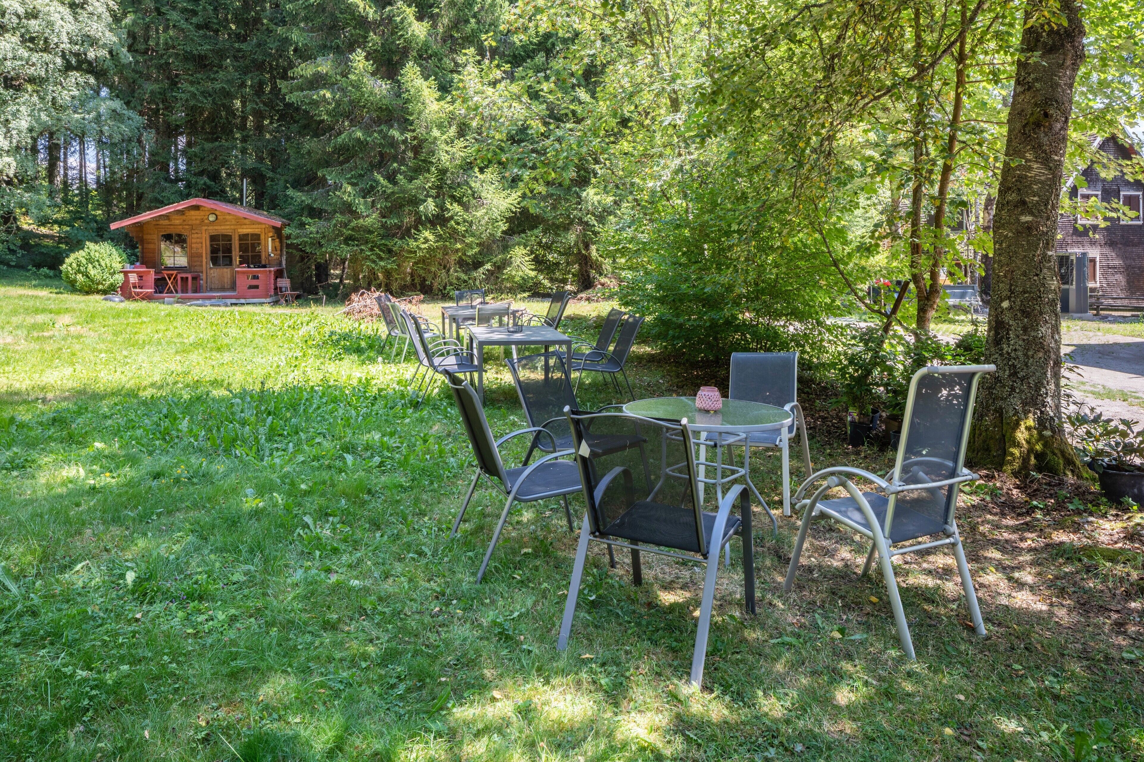 Un espace extérieur aménagé avec des chaises et des tables en métal, entouré d’une végétation luxuriante et agrémenté d’un petit bâtiment en bois en arrière-plan.