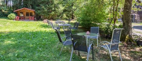 Un espace extérieur aménagé avec des chaises et des tables en métal, entouré d’une végétation luxuriante et agrémenté d’un petit bâtiment en bois en arrière-plan.