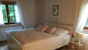Un dormitori amb un llit, una tauleta de nit amb una làmpada, una finestra amb cortines i un quadre penjat a la paret.