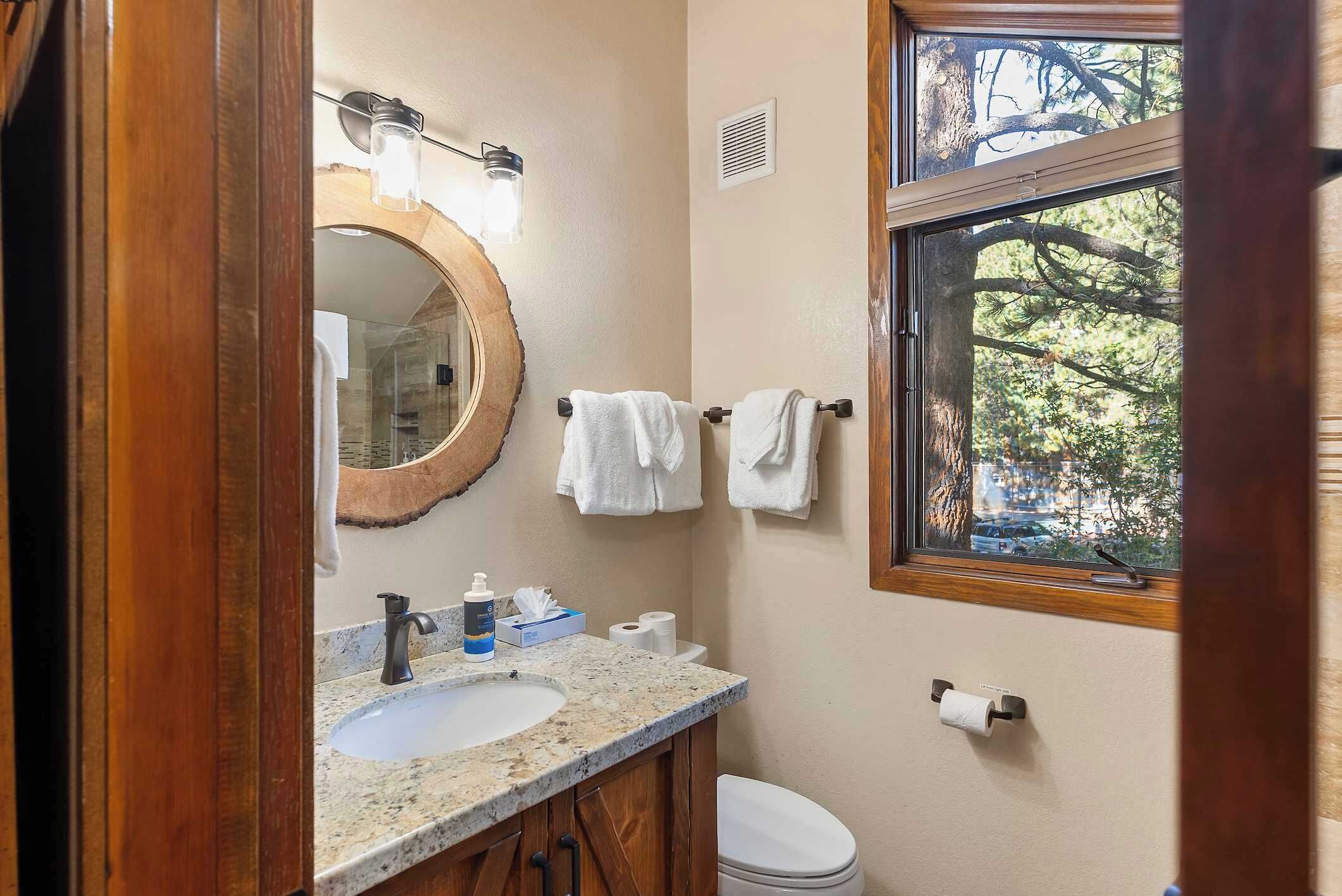 A bathroom with a sink, mirror, towel rack, toilet, and a window with a view of trees.