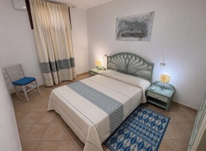 Ein Schlafzimmer mit Bett, Sessel, Nachttisch, Lampe, Wandbild und einem gemusterten Teppich.