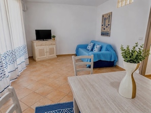 Ein kleiner, möblierter Raum mit Fernseher auf einem Schrank, einem blauen Sofa, einem weißen Sessel und einem Tisch mit einer Vase.