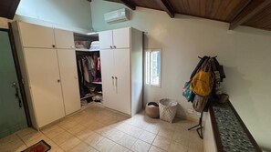 Ein Zimmer mit Schrank, kleinem Fenster, Mülleimer und einer Bank mit Taschen.
