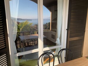 A balcony with a view of a body of water and distant land, a table, chairs, and a kitchen area. - 180° View of Lake Maggiore and the Alps (Meina)