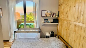 Un dormitori amb llit, finestra amb cortines, vista a zones verdes a l'exterior, una estanteria amb llibres i un radiador.
