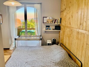 A bedroom with a bed, a window with curtains, a view of greenery outside, a shelf with books, and a radiator. - 180° View of Lake Maggiore and the Alps (Meina)