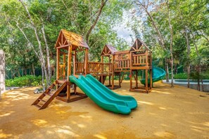 Ein Spielplatz mit hölzernen Spielgeräten, darunter eine Rutsche und Kletterbereiche, umgeben von Bäumen und Grünflächen.