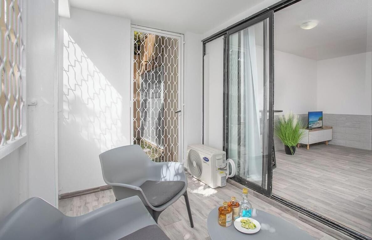 A modern living room with a sliding glass door leading to a balcony, a grey sofa, a small table with bottles, and a plant on the balcony.