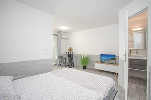 A modern hotel room with a bed, a TV, a small table with chairs, a potted plant, and a bathroom visible through an open door. - The Jewel of Terreville. T1. 5 minutes from the beaches. (Schœlcher)