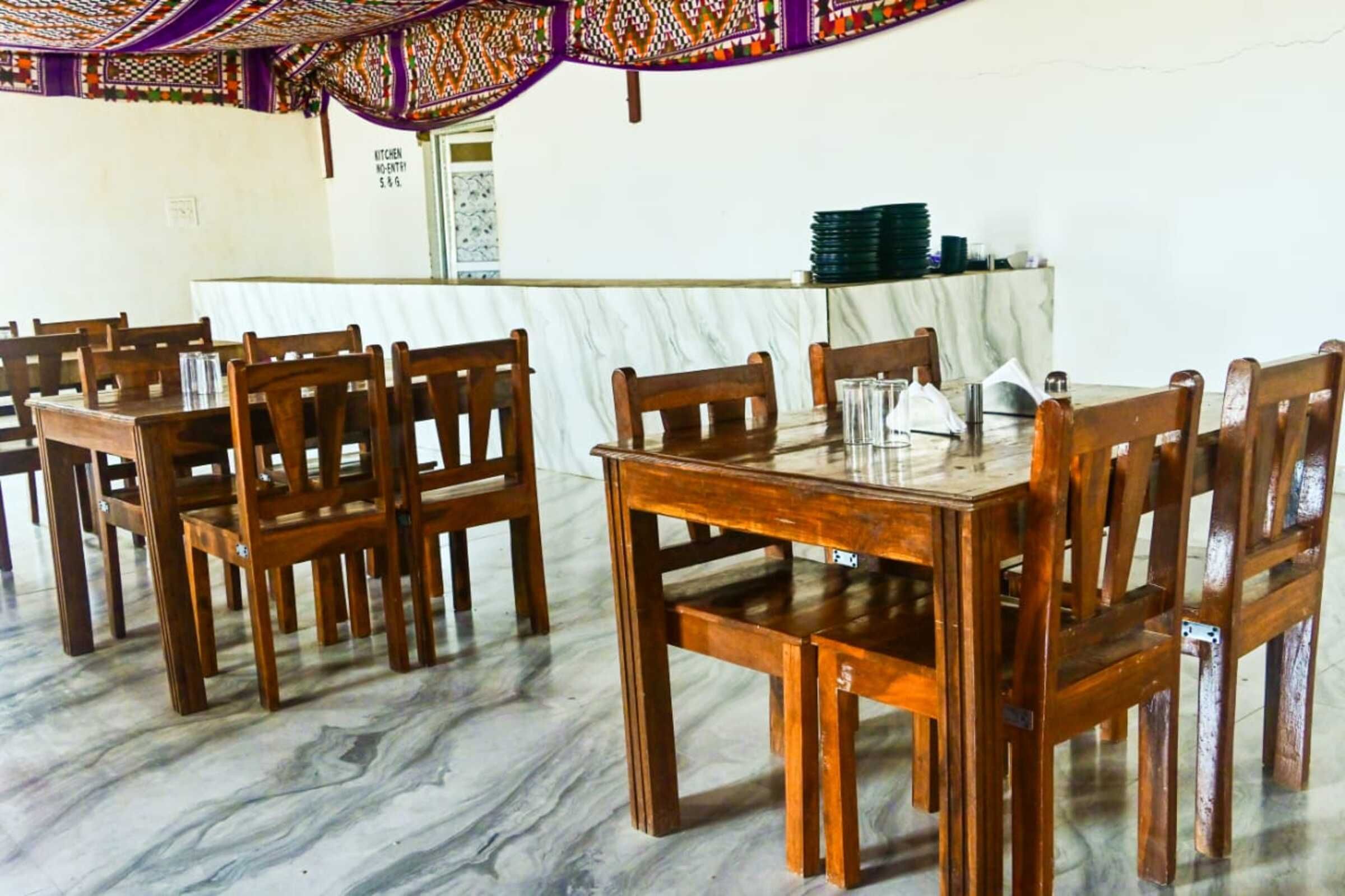 A dining area with wooden tables and chairs, a marble floor, and a kitchen area in the background.