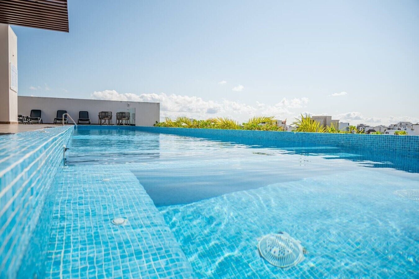 A rooftop infinity pool with clear blue water, surrounded by a white wall and a row of black chairs.