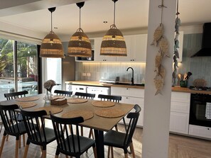 A modern kitchen with a dining area, featuring a large table with chairs, pendant lights, and a view of the outdoors.