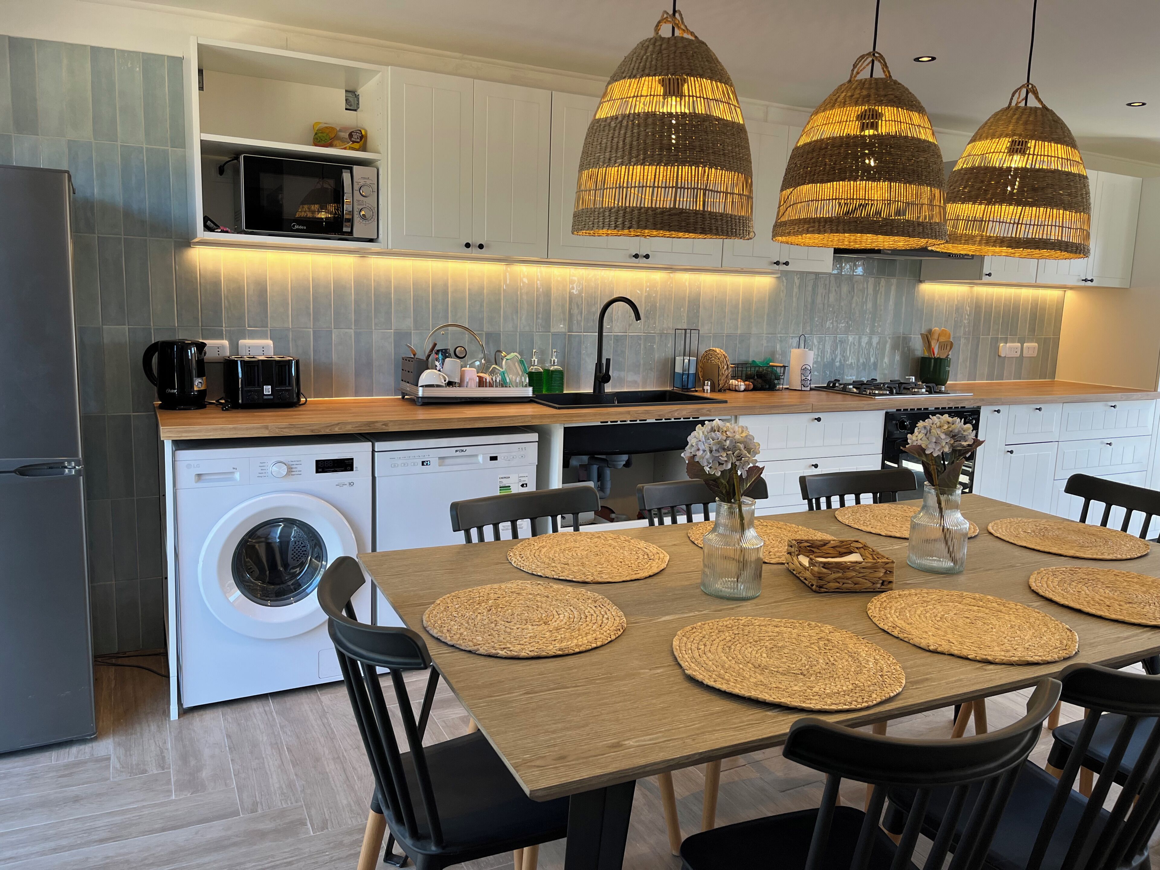 A modern kitchen with built-in appliances, a dining area with a wooden table and chairs, and hanging wicker pendant lights.