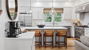 Una cocina moderna con armarios blancos, una isla grande con taburetes de bar, una cafetera y vistas al exterior.