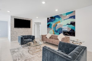 Een moderne woonkamer met een groot abstract schilderij, een flatscreen televisie en een bank met twee decoratieve kussens.