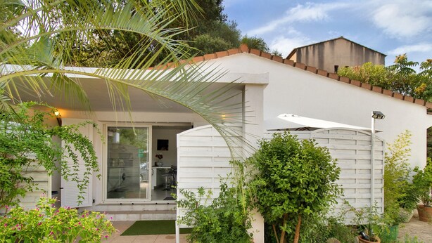 A modern house with a covered patio, a visible kitchen area, and a variety of plants.