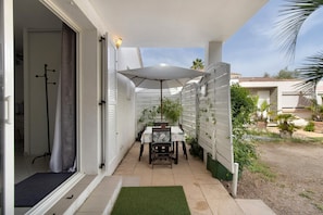 Une terrasse avec une table et des chaises, un parasol blanc et un petit jardin.